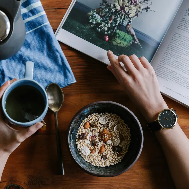 Frühstückstisch von oben: eine Tasse Tee, eine Schale Müsli und eine aufgeschlagene Zeitschrift